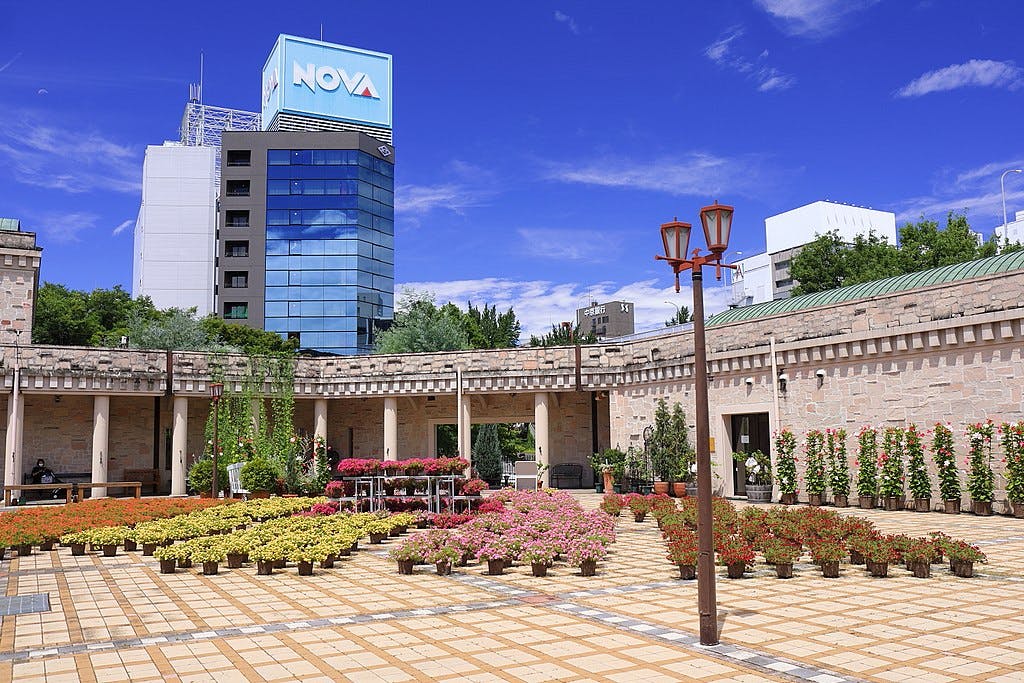 A sunny plaza with flowerbeds arranged in patterns, surrounded by a stone colonnade. Modern office buildings, including one with a large "NOVA" sign, rise in the background against a blue sky.