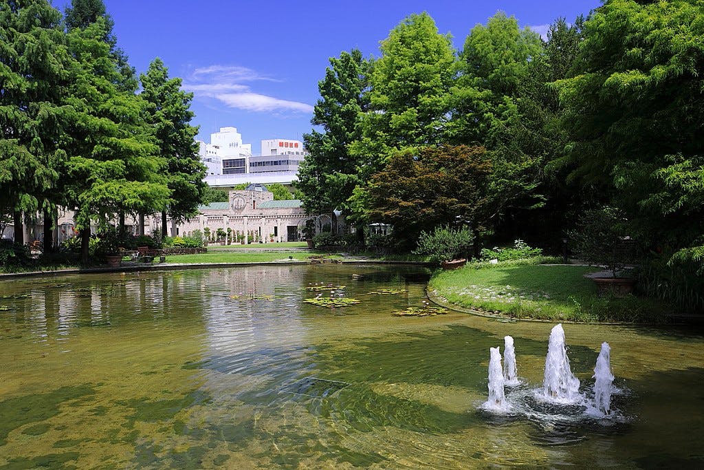 A tranquil park scene with a pond, water fountain, green trees, and lily pads. In the background, there are historic stone buildings and modern city structures under a clear blue sky.