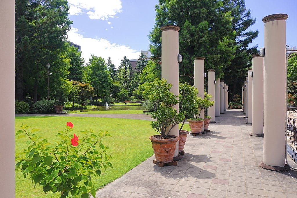 A landscaped garden with green grass, trees, potted plants, and a row of tall white columns along a tiled walkway under a bright blue sky. A red flower blooms in the foreground.