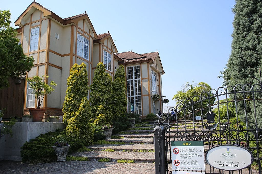 A large, light-colored house with brown trim, tall windows, and a steep roof sits behind a black metal gate. Stone steps lead up to the entrance, surrounded by manicured shrubs and trees on a sunny day.