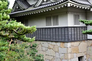 Traditional Japanese building with white walls, tiled roof, and wooden lattice windows, set on a stone foundation and surrounded by lush green trees and shrubs.