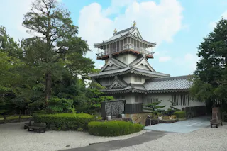 A traditional Japanese castle with white walls and gray tiled roofs, surrounded by trees and a gravel courtyard under a partly cloudy sky. Benches and a stone monument are visible in the foreground.