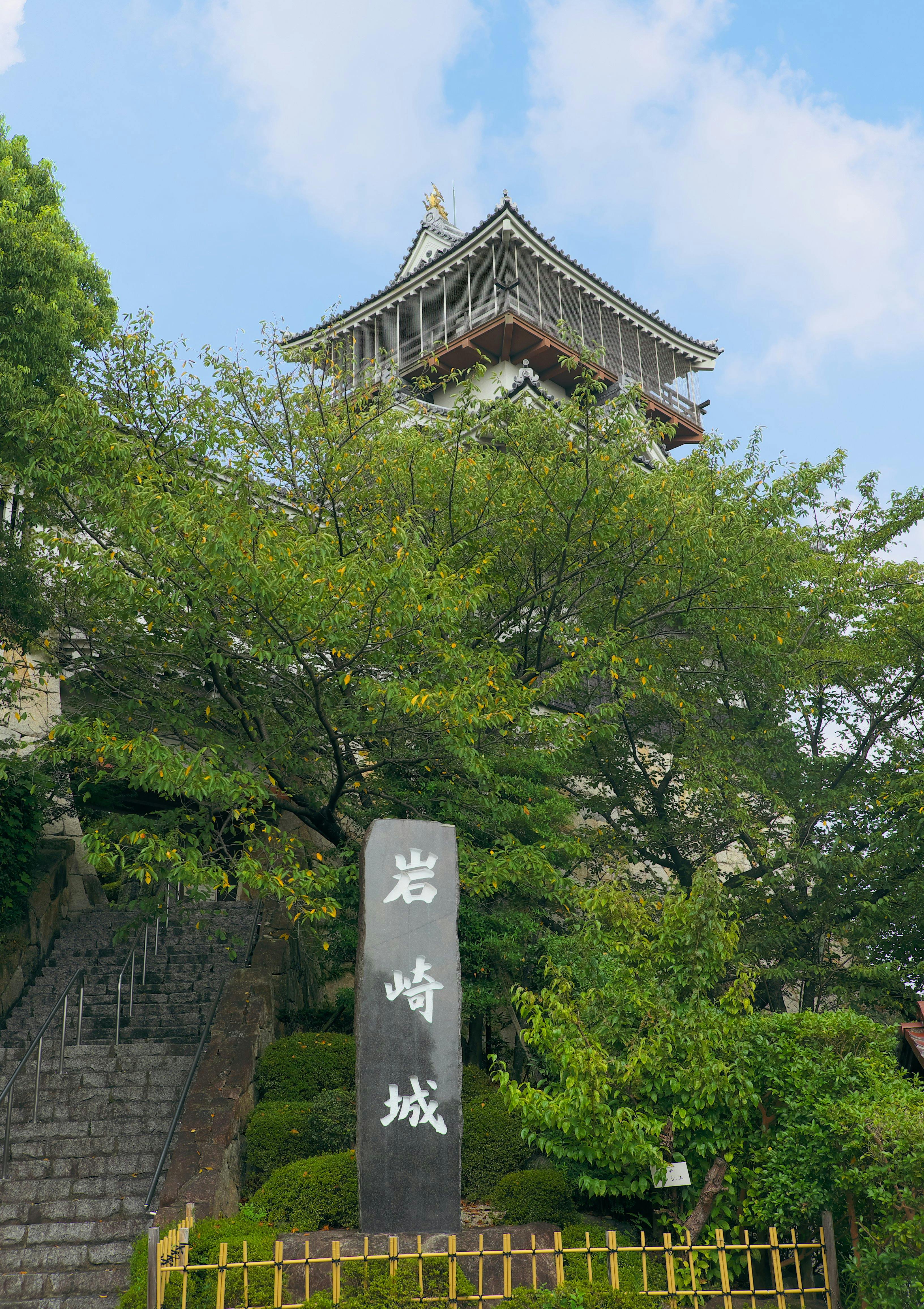 A traditional Japanese castle tower rises behind green trees, with a stone monument featuring Japanese inscriptions in the foreground and a stone staircase to the left, all under a partly cloudy sky.