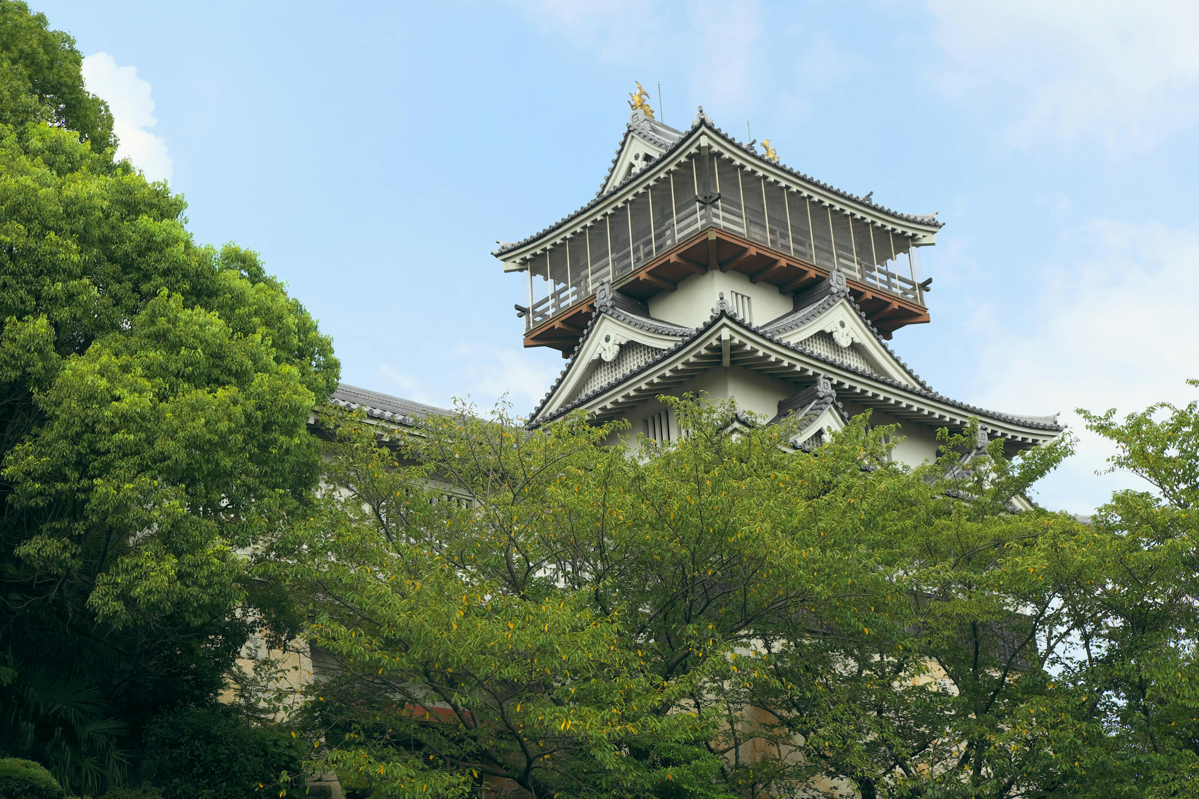 A traditional Japanese castle with ornate rooflines rises above green trees, set against a partly cloudy blue sky.