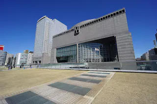 A large, modern gray building with geometric shapes and glass windows stands under a clear blue sky. The area in front features a wide walkway and a neatly trimmed grass lawn with several people walking nearby.