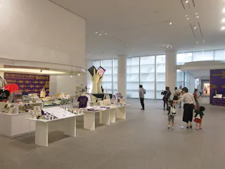 Spacious modern museum lobby with large windows, several people walking or standing, and a shop area displaying various goods and souvenirs on white tables. Purple signs and decor are visible in the background.