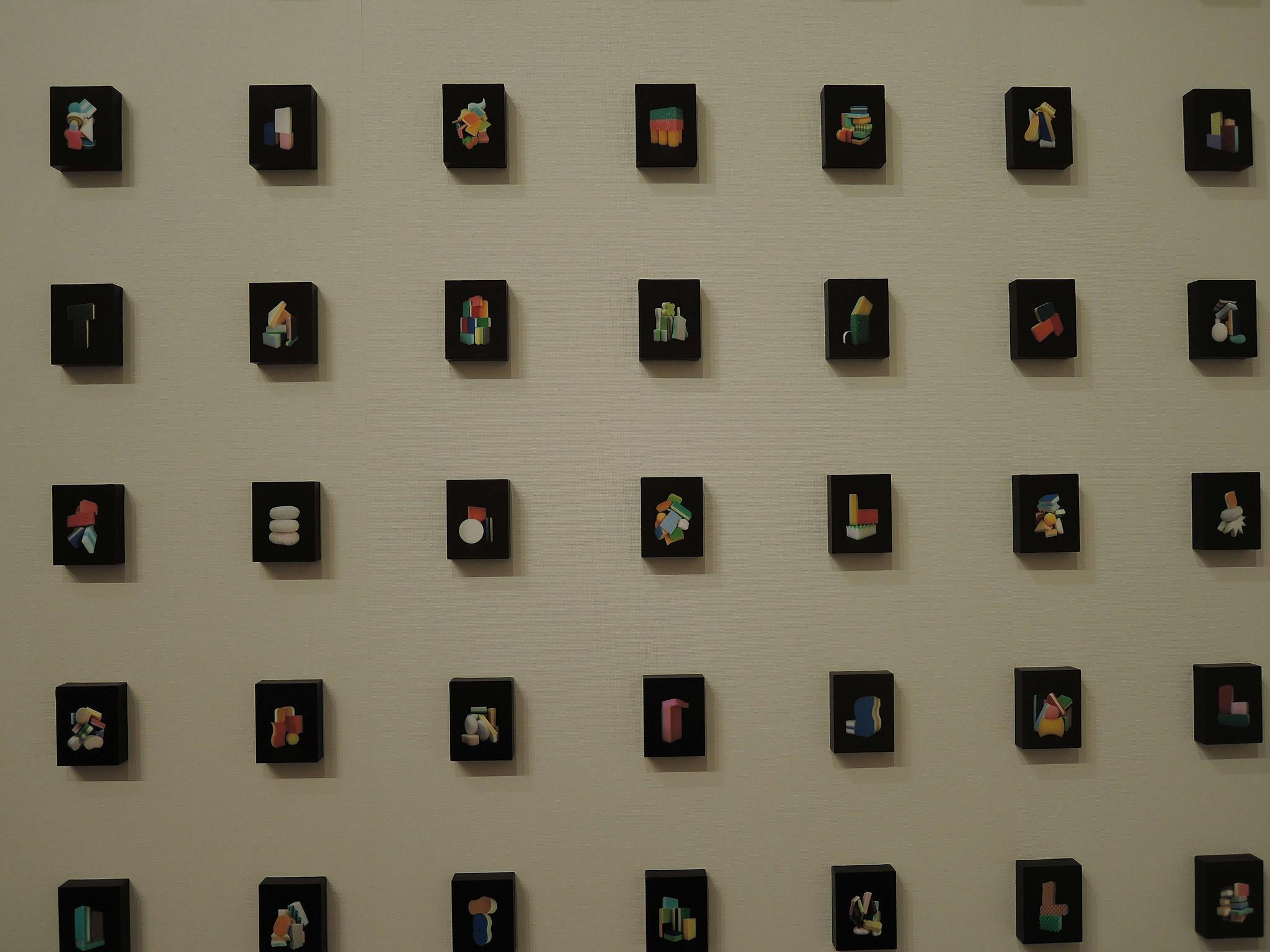 A grid of small black square plaques is mounted on a beige wall, each displaying a unique, colorful abstract 3D shape in pastel colors. The arrangement is orderly and evenly spaced.