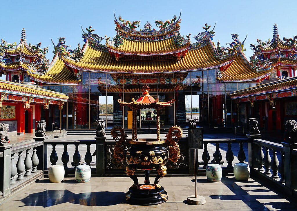 A colorful, ornate temple with detailed carvings and golden roofs under a clear blue sky. In the foreground is an elaborate incense burner, surrounded by stone railings and decorative jars.