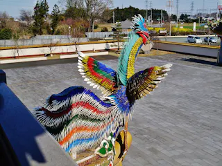 A large, colorful phoenix statue with outstretched wings stands in an open, paved courtyard. The detailed sculpture features vibrant blues, reds, greens, and yellows, with trees and fields in the background.