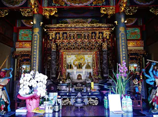 Ornate altar in a Chinese temple, decorated with intricate gold details, statues, colorful columns, floral offerings, drinks, and ceremonial objects, with vivid artwork on the walls and ceiling.