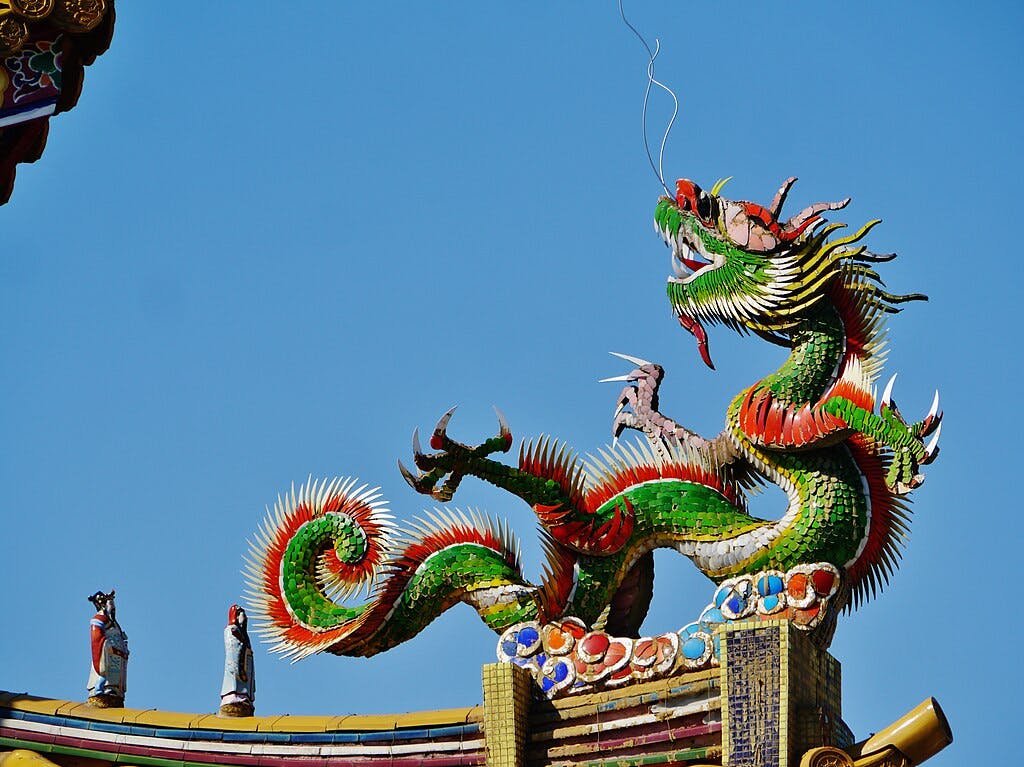 A colorful dragon sculpture with red, green, and yellow details sits atop a traditional temple roof against a clear blue sky. There are also small human figures on the roof beside the dragon.
