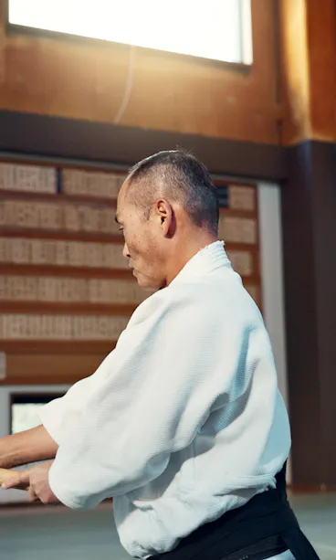 Samurai Class An older man in a martial arts uniform practices with a wooden sword in a dojo, standing on a mat in front of wooden plaques and a window that lets in natural light.