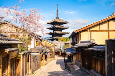 A traditional street in Kyoto, Japan, with wooden houses on both sides, cherry blossoms in bloom, and a five-story pagoda standing tall in the background under a blue sky.