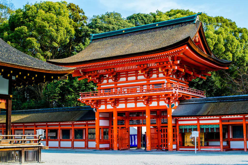 A traditional Japanese shrine with vibrant red wooden structures and a large torii gate. The building features a curved roof and intricate architectural details, set against a backdrop of lush green trees under a clear blue sky.