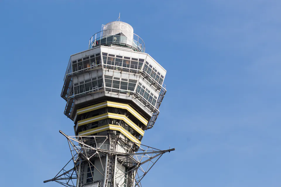 Close-up of a tall, hexagonal observation tower with large glass windows and a metallic framework, set against a clear blue sky. The structure has a modern design with several levels and antennas on top.
