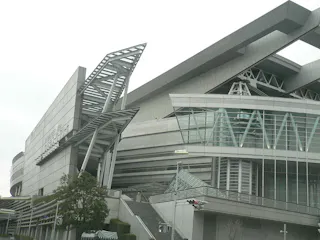 A modern building with angular, futuristic architecture, featuring large glass windows and metal structures, under a cloudy sky. Some greenery and a traffic light are visible in the foreground.