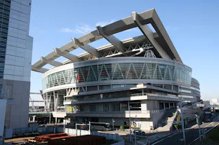A large modern sports arena with a circular glass facade and geometric roof supports, surrounded by roads and buildings, under a clear blue sky. Buses and cars are parked nearby.