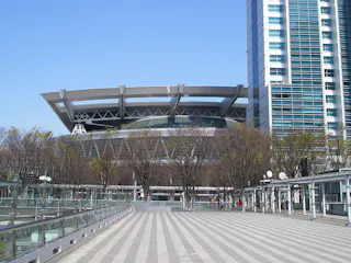 A modern stadium with a distinctive, curved roof behind leafless trees and next to a tall glass building, with a wide, empty plaza in the foreground under a clear blue sky.