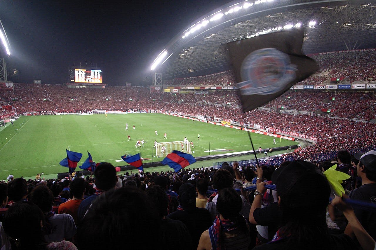 A large, crowded stadium at night with fans waving flags and watching a soccer match. The field is brightly lit, and the stands are filled with spectators wearing red and blue colors.