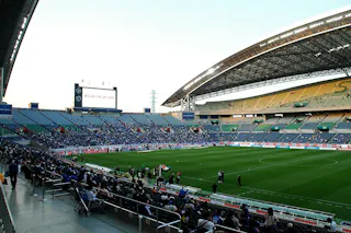 A large, modern football stadium with a partially covered roof, filled with fans seated in the stands. The playing field is green and there is a large screen displaying information at one end of the stadium.