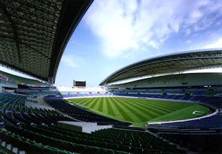 A wide-angle view of an empty modern stadium with green grass, striped patterns on the field, rows of blue and green seats, and a large curved roof under a partly cloudy sky.