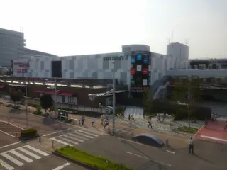 A modern shopping mall named Cocoon 3 with a white checkered facade, colorful signage, and people walking on the sidewalks and crosswalk in front of the building on a sunny day.