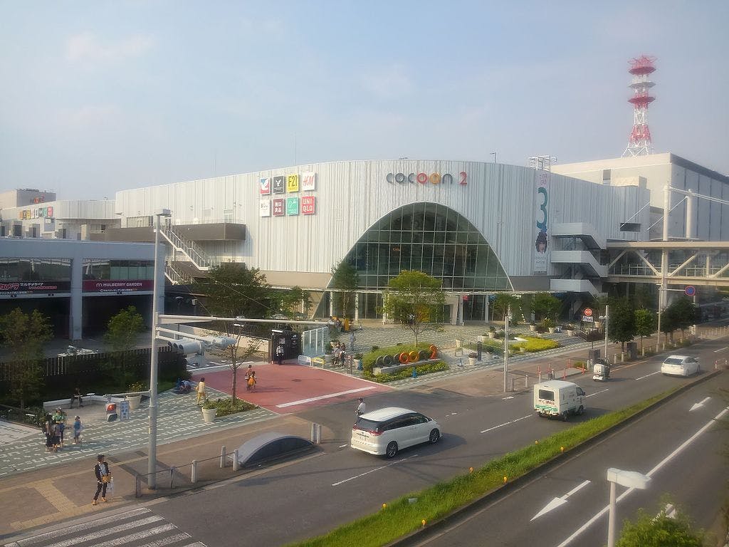 Large modern shopping mall with a white exterior and arched glass entrance, called Cocoon 2. Cars and people are visible outside, and a red and white tower stands in the background. Trees and crosswalks surround the area.