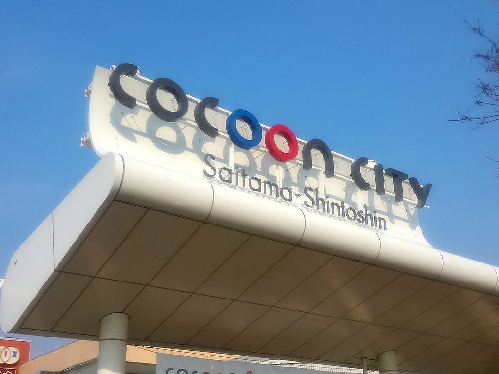 A sign reading "Cocoon City Saitama-Shintoshin" is mounted on a modern white structure against a clear blue sky. The words "Cocoon City" use stylized letters with interlinked O's.