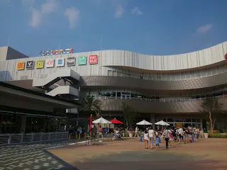 A modern shopping mall with multiple brand signs, including UNIQLO and H&M, above the entrance. People walk and gather in the open plaza below, some under white umbrellas, on a sunny day.