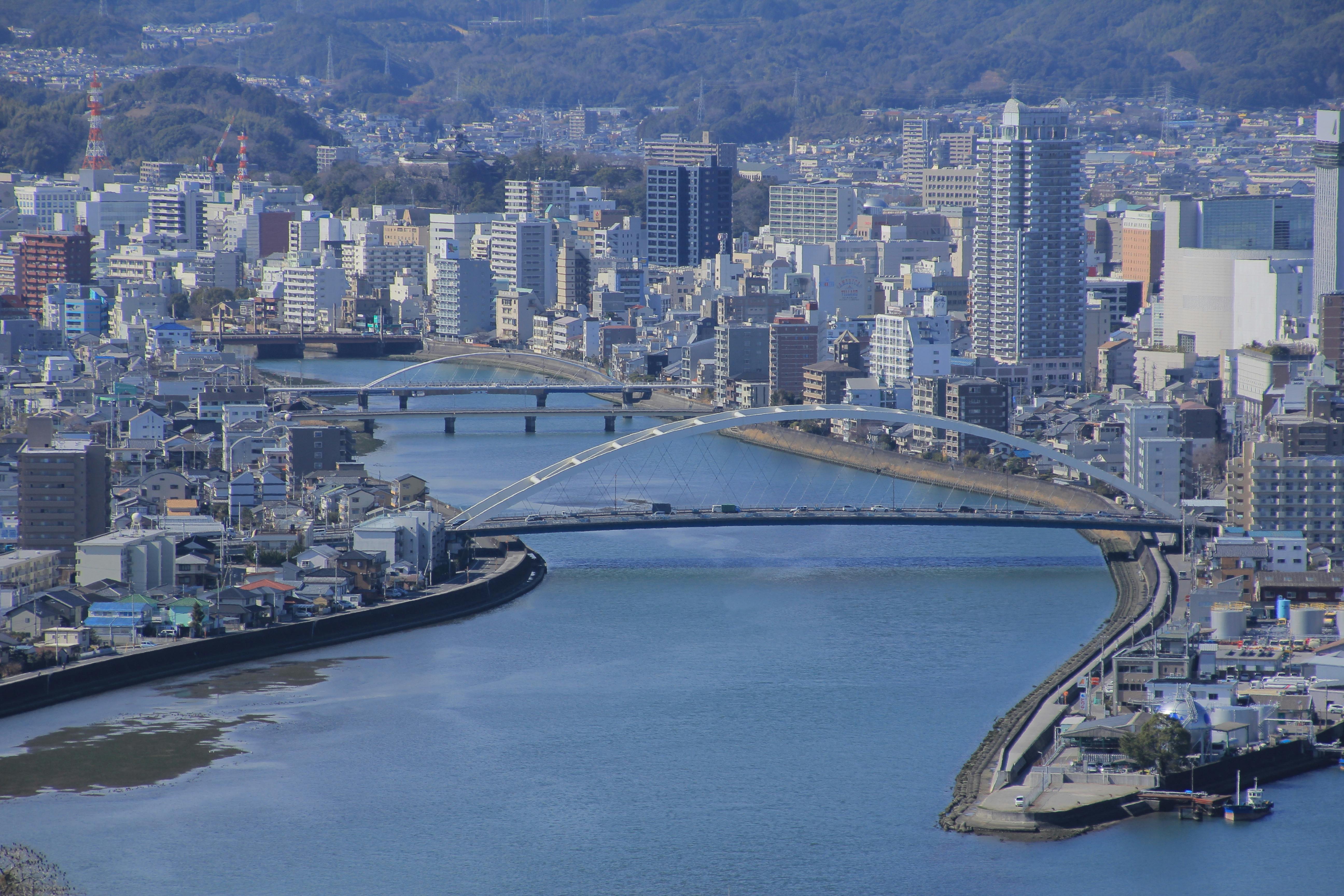 A cityscape featuring a wide river with two modern arch bridges, lined by buildings and high-rises on both sides, and tree-covered hills in the background under a clear sky.