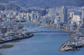 A cityscape featuring a wide river with two modern arch bridges, lined by buildings and high-rises on both sides, and tree-covered hills in the background under a clear sky.