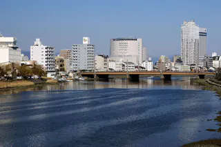 A calm river flows under a bridge, with modern mid-rise and high-rise buildings lining the riverbanks under a clear blue sky.