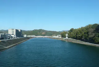 A wide river with blue water flows between concrete embankments. On the left are mid-rise buildings; on the right, dense green trees. Hills and a clear sky are visible in the background. A bridge crosses the river ahead.