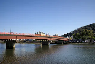 A red pedestrian bridge spans over a calm river with buildings and trees in the background under a clear blue sky.