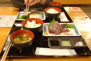 A Japanese meal set on a wooden table featuring bowls of white rice, miso soup, slices of seared fish with garnish, dipping sauces, and chopsticks; a hand holds chopsticks over one tray.