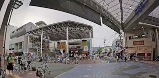 A busy Japanese shopping street with a covered walkway, parked bicycles, people walking, and colorful banners and signs on buildings under a large metal and glass canopy.