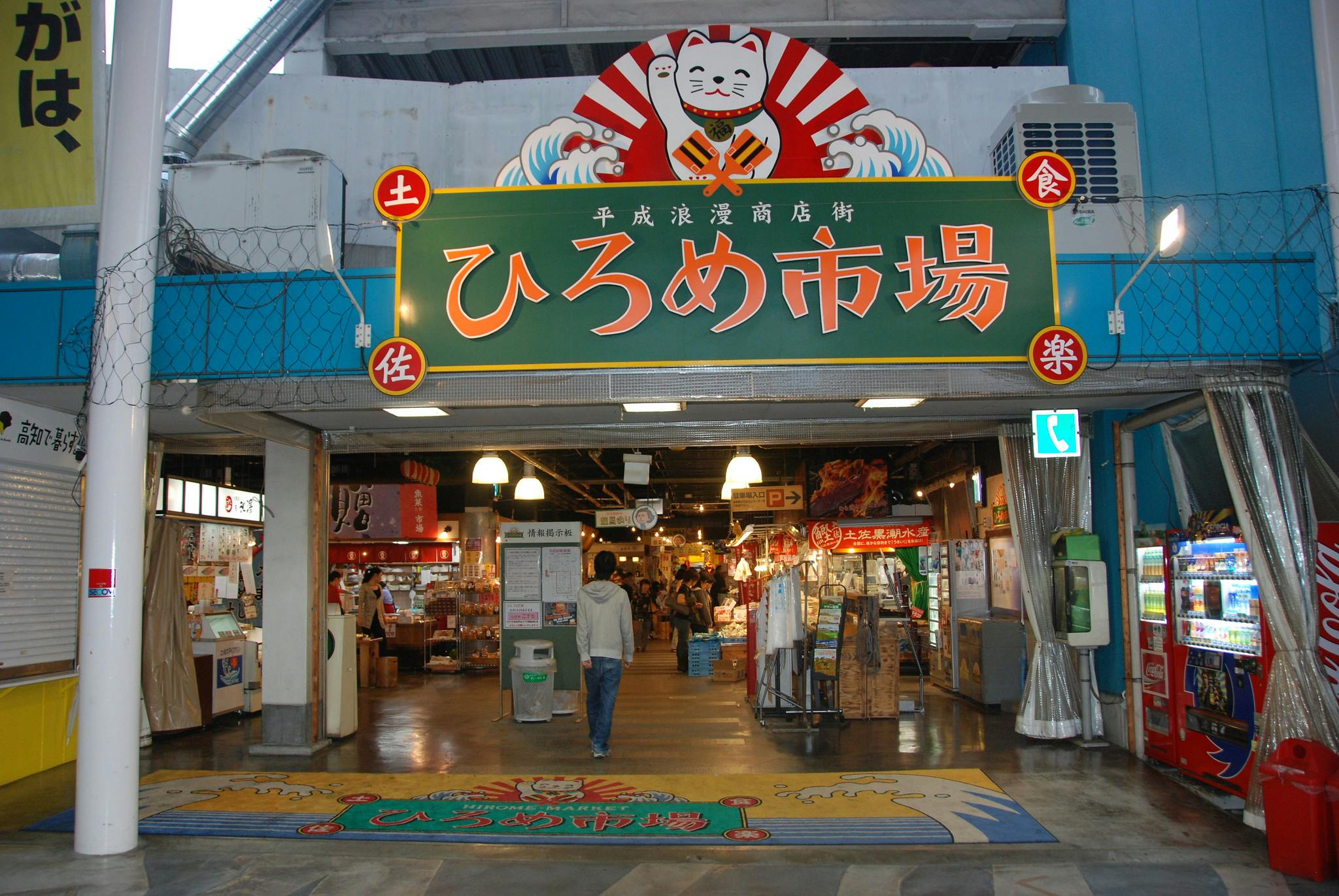 Entrance to a Japanese indoor market, decorated with a large sign in Japanese, a lucky cat figure, and colorful banners. Stalls with lights and food displays are visible inside, with one person walking in.