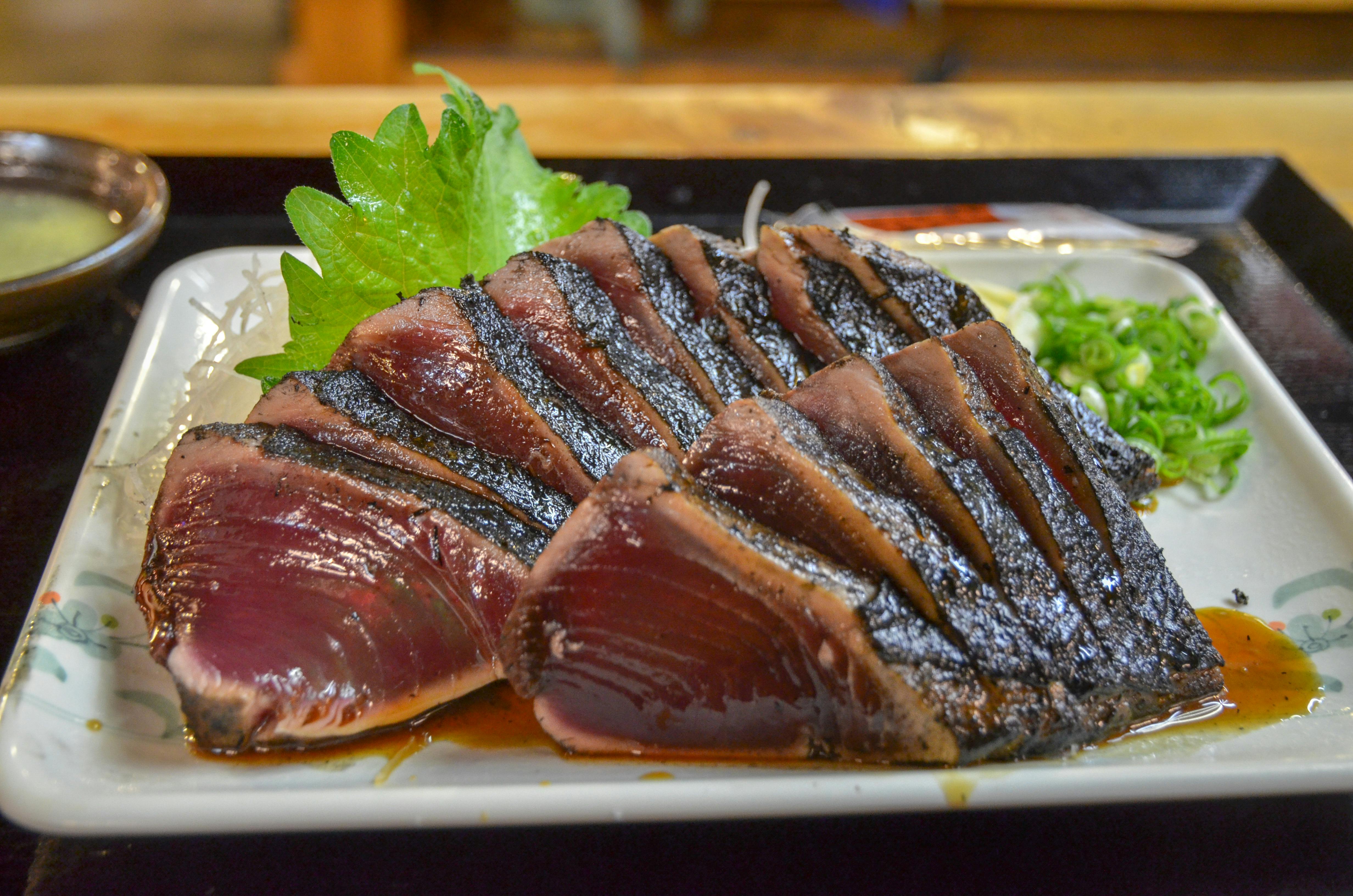 A plate of seared tuna sashimi slices, arranged neatly and garnished with green shiso leaves and chopped scallions, sits on a white rectangular dish with soy-based sauce.