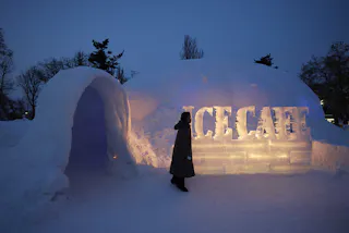 A person in a coat stands outside an illuminated ice structure labeled "ICE CAFE" at dusk, with snow and trees in the background.
