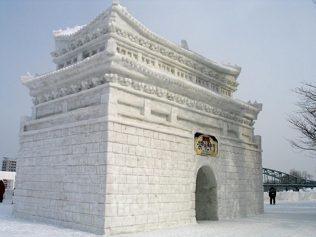 A large, detailed snow sculpture resembling a traditional East Asian gate or fortress, with arched entryway and tiered roof, stands outdoors on a snowy day. A few people are visible for scale.