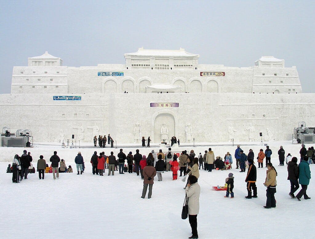 A large crowd gathers in front of an enormous, intricately carved snow sculpture resembling a palace or fortress, with people scattered across a snowy field under a cloudy sky.