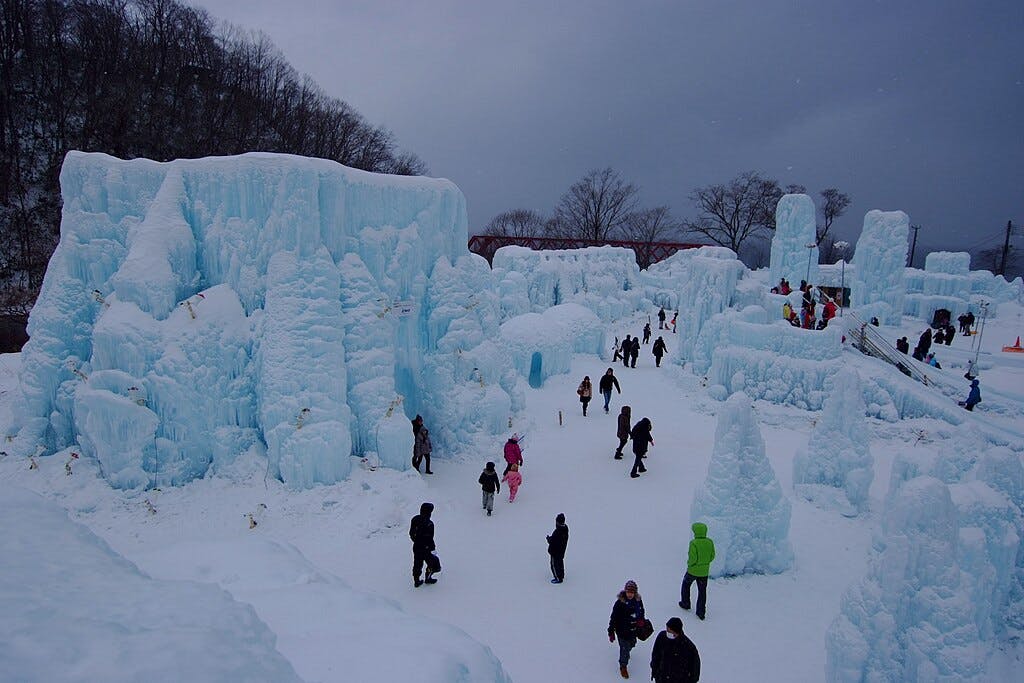 People walk among large, sculpted ice formations and snow under a cloudy sky in a winter landscape surrounded by bare trees.
