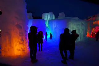People stand inside an ice cave at night, surrounded by large ice walls illuminated with colorful lights in shades of orange, blue, green, and red, creating a vibrant and magical atmosphere.