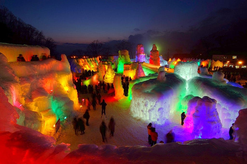 An outdoor ice festival at dusk features large, colorful ice sculptures illuminated by vibrant lights, with crowds of people walking among the glowing structures under a dark blue sky.