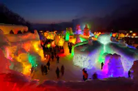 An outdoor ice festival at dusk features large, colorful ice sculptures illuminated by vibrant lights, with crowds of people walking among the glowing structures under a dark blue sky.