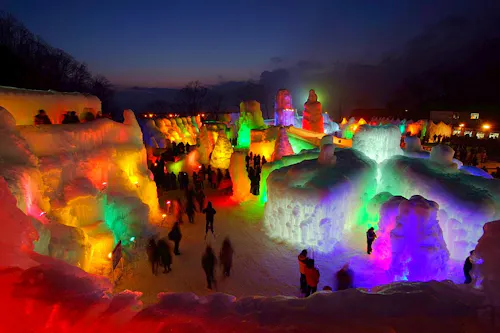 An outdoor ice festival at dusk features large, colorful ice sculptures illuminated by vibrant lights, with crowds of people walking among the glowing structures under a dark blue sky.