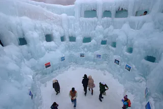 A group of people stand inside a circular ice structure with thick icy walls and windows. Pictures are displayed on the walls, and the ground is covered in snow. The scene appears cold and wintry.