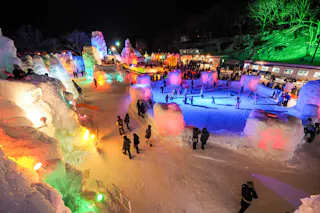 A vibrant nighttime scene at a winter festival with people walking among large, colorful ice sculptures illuminated by lights, and others skating on an ice rink surrounded by snow and glowing displays.