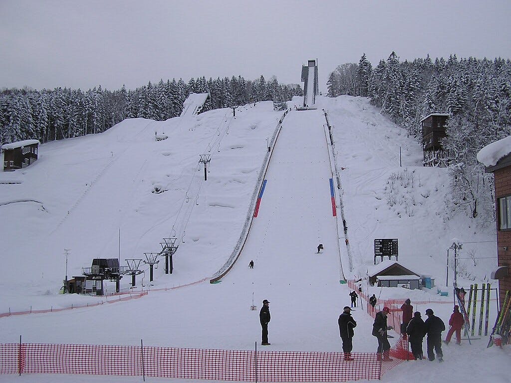 A snowy ski jump hill surrounded by trees, with people standing at the bottom and a few on the slope. Buildings and a ski lift are visible to the sides, under an overcast sky.