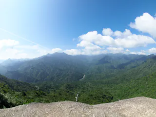 A panoramic view of lush green mountains under a bright blue sky with scattered white clouds, seen from a rocky vantage point in the foreground.